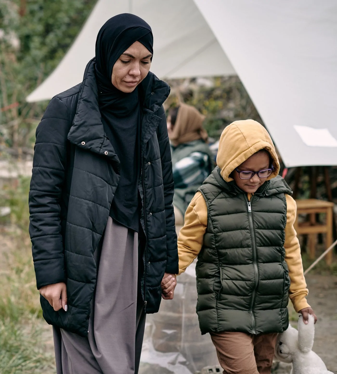 A mother and her daughter walk hand in hand through a camp of tents